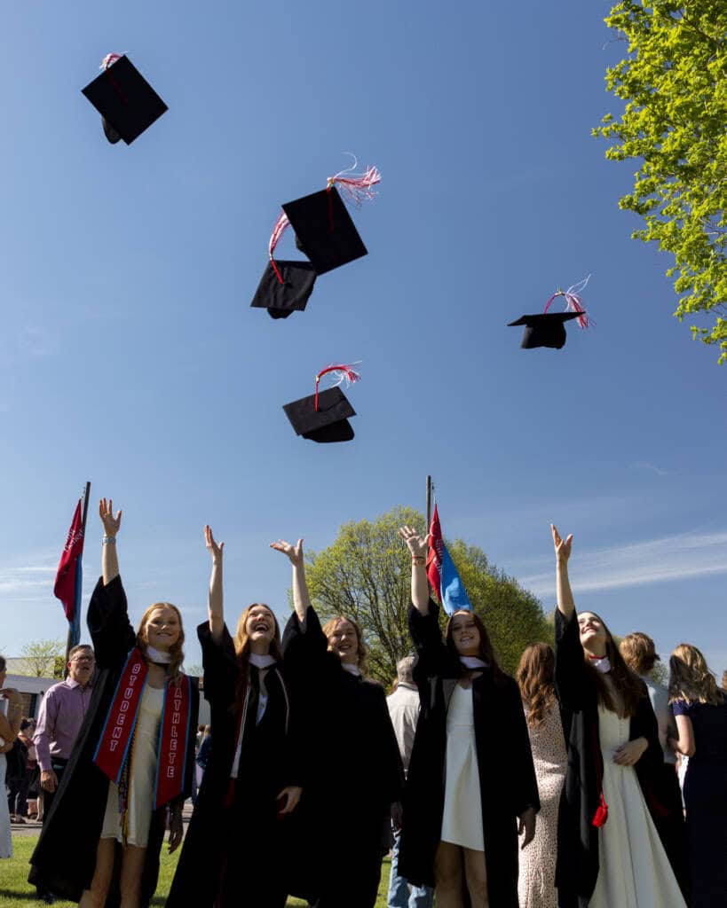 Five graduates in caps and gowns joyfully toss their caps into the air during an outdoor graduation ceremony on a sunny day, with a crowd and flags in the background.
