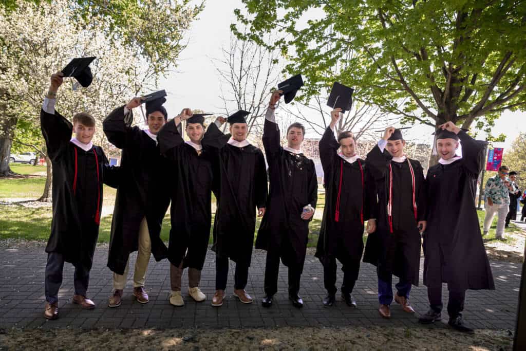 Seven graduates in black caps and gowns stand outdoors, smiling and holding their caps up. Trees with green leaves and a sunny sky are in the background, giving the scene a celebratory atmosphere.