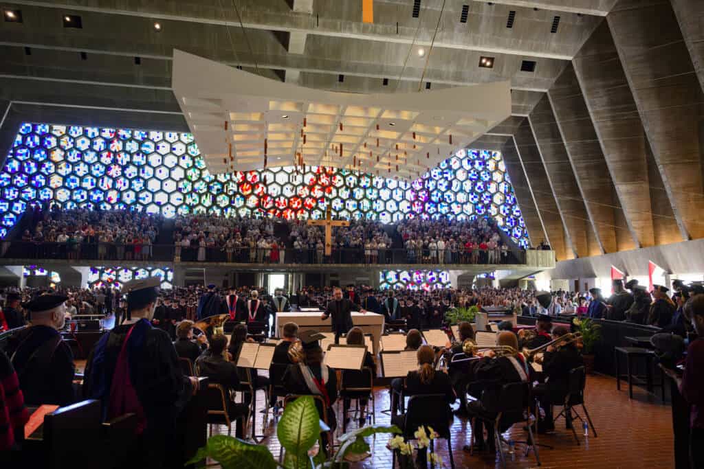 A large audience fills a modern church with colorful stained glass windows, as a conductor leads an orchestra. People in academic robes are seated in the foreground, and many attendees watch from a balcony.