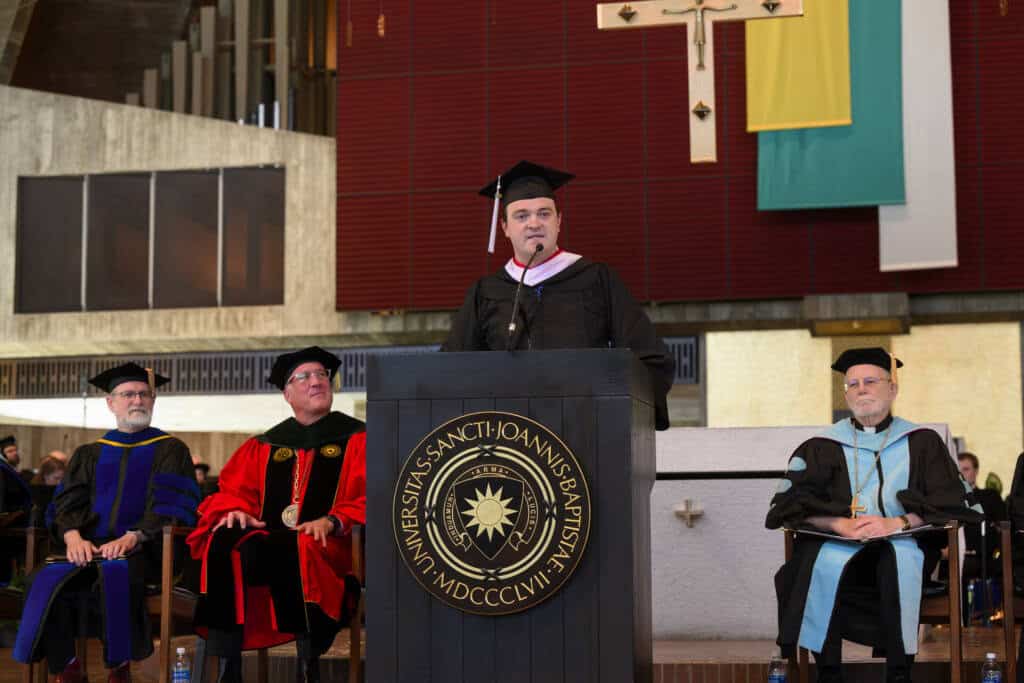 A man in academic regalia speaks at a podium during a graduation ceremony, with three other officials seated behind him on stage in similar academic attire.