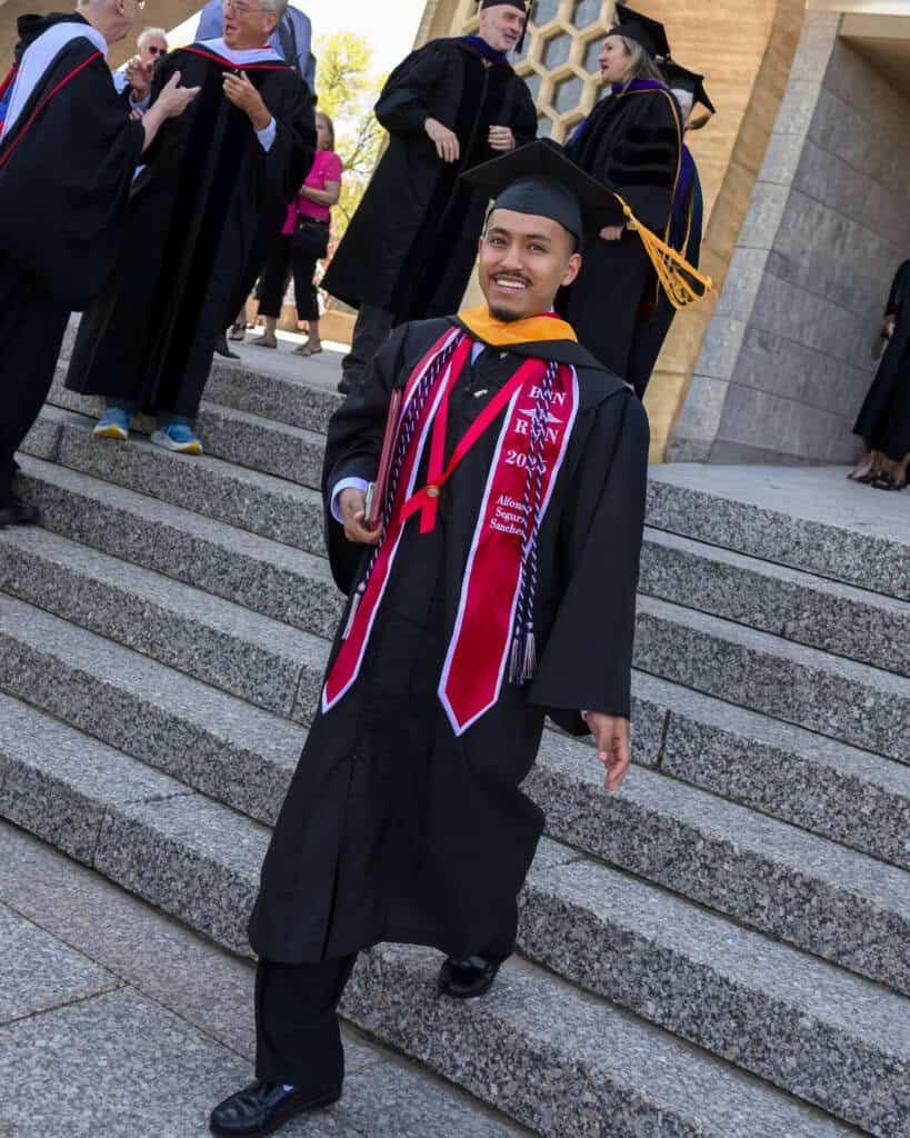 A smiling graduate in a cap and gown walks down outdoor steps, wearing a red and white stole and holding a diploma, with other graduates and faculty in robes in the background.