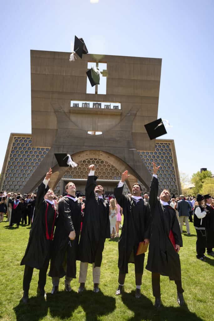 Five graduates in black gowns throw their caps into the air in celebration outside a modern building, surrounded by other graduates and attendees on a sunny day.
