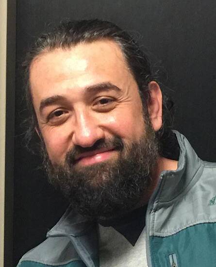 A person with long-brown hair and a full beard, smiling at the camera. They are wearing a green and gray jacket over a light-colored shirt. The background is a dark-colored wall.
