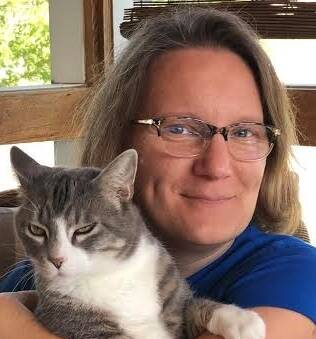 A woman with glasses, wearing a blue shirt, is sitting on a striped couch and holding a large gray and white cat in her arms. The woman is smiling, and the cat looks relaxed. There is a window with greenery visible in the background.