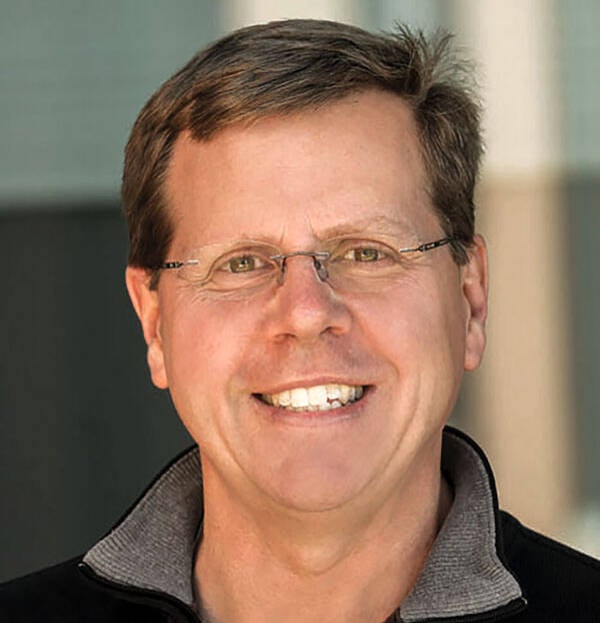 A smiling person with short brown hair and glasses. They are wearing a black zip-up jacket with a gray collar. The background appears to be an indoor setting with blurred features.