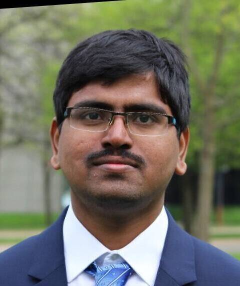 A person with short dark hair and glasses is shown outdoors, wearing a blue suit and a light blue tie with a subtle pattern. The background consists of green trees and grass with a building partly visible.
