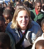A smiling person with light hair wearing a denim jacket is surrounded by a group of children. The children are wearing school uniforms and are gathered closely together outdoors.