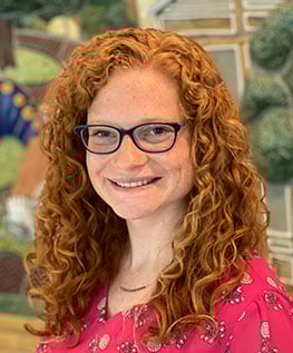 A woman with long curly red hair and glasses, wearing a pink blouse with a floral pattern, smiles at the camera. The background appears to be a colorful, abstract mural, featuring various shapes and textures.