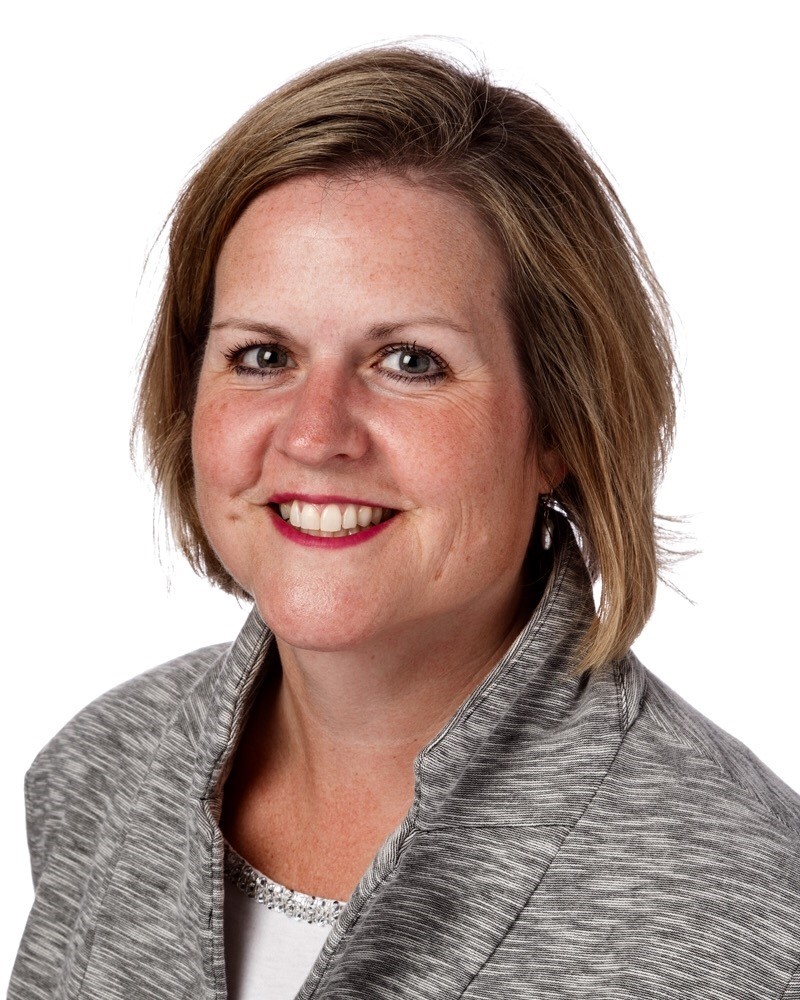 A woman with short, light brown hair smiles warmly at the camera. She is wearing a gray jacket over a white top. The background is plain white, making her the focal point of the image.