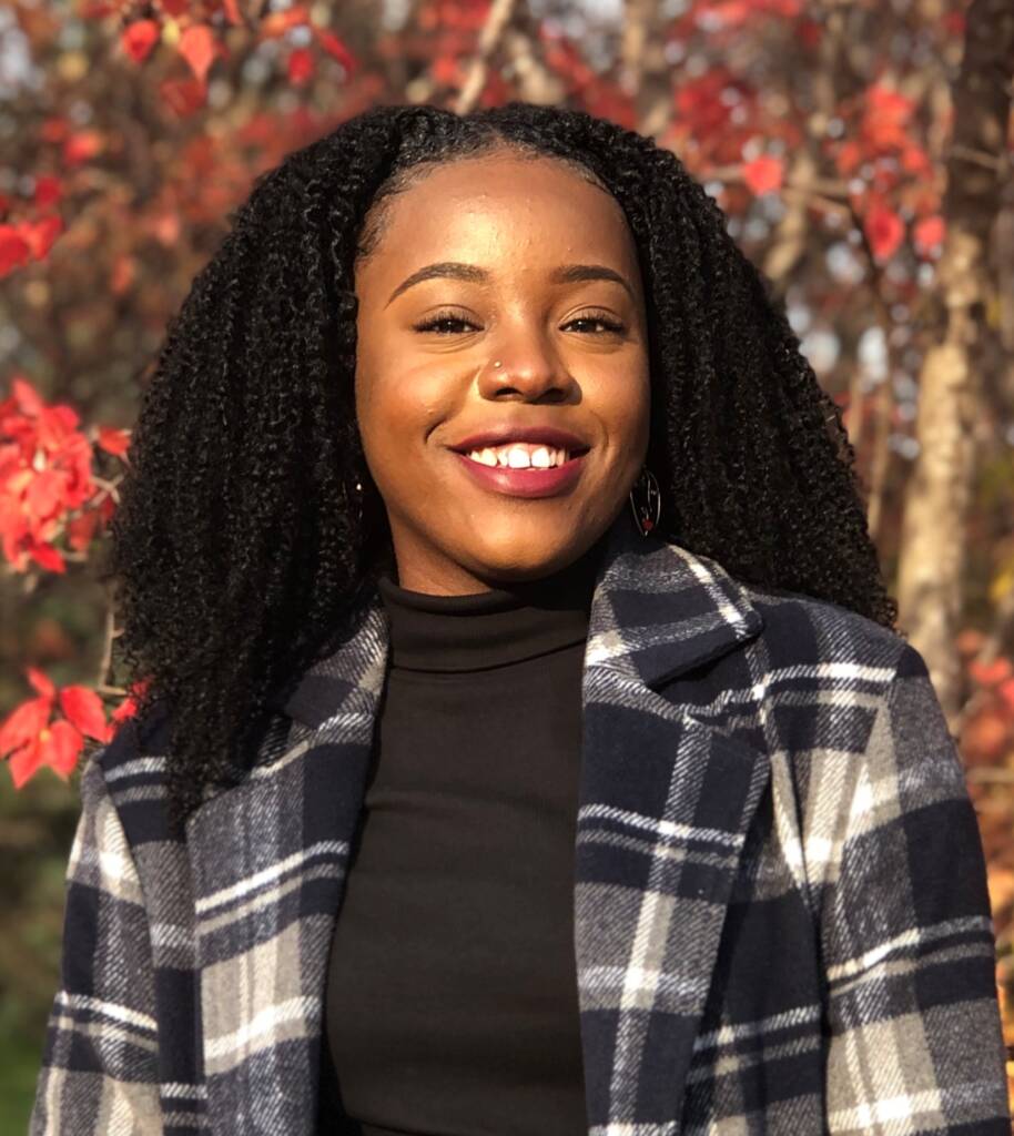 A woman with long curly hair smiles at the camera. She is wearing a black turtleneck and a blue and white plaid jacket. The background contains trees with red autumn leaves.