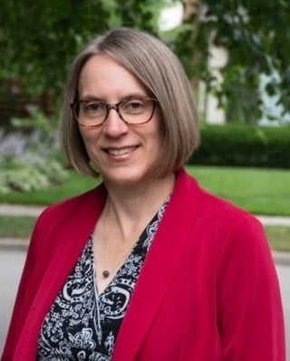 A person with shoulder-length gray hair and glasses is smiling. They are wearing a patterned black and white top and a red blazer. The background shows greenery with blurred foliage and a lawn.