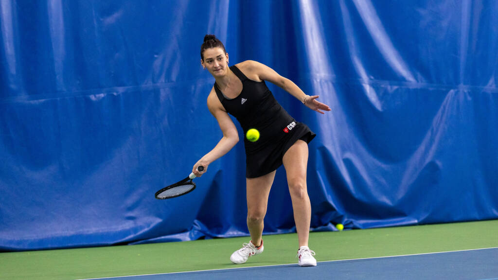 A tennis player in a black outfit lunges to hit a forehand shot. She is on a blue indoor court with a blue curtain in the background.