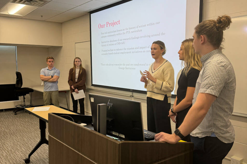 Five individuals stand in a classroom giving a presentation. Three women and two men are present, with one woman speaking while the others listen. A projection screen displays the title "Our Project" with text below. Desks and chairs are in the background.