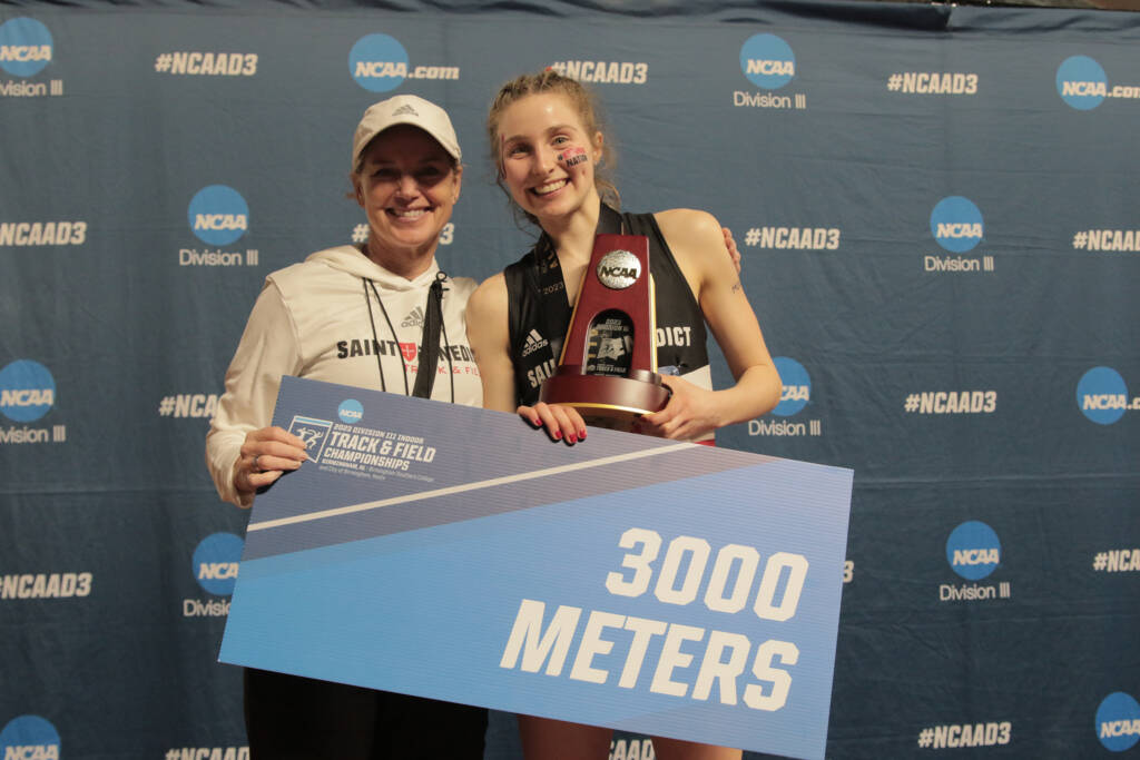 Two people pose happily in front of a blue NCAA Division II backdrop. One holds a championship trophy, and the other holds a large card that reads "3000 METERS" and "TRACK & FIELD CHAMPIONSHIPS.