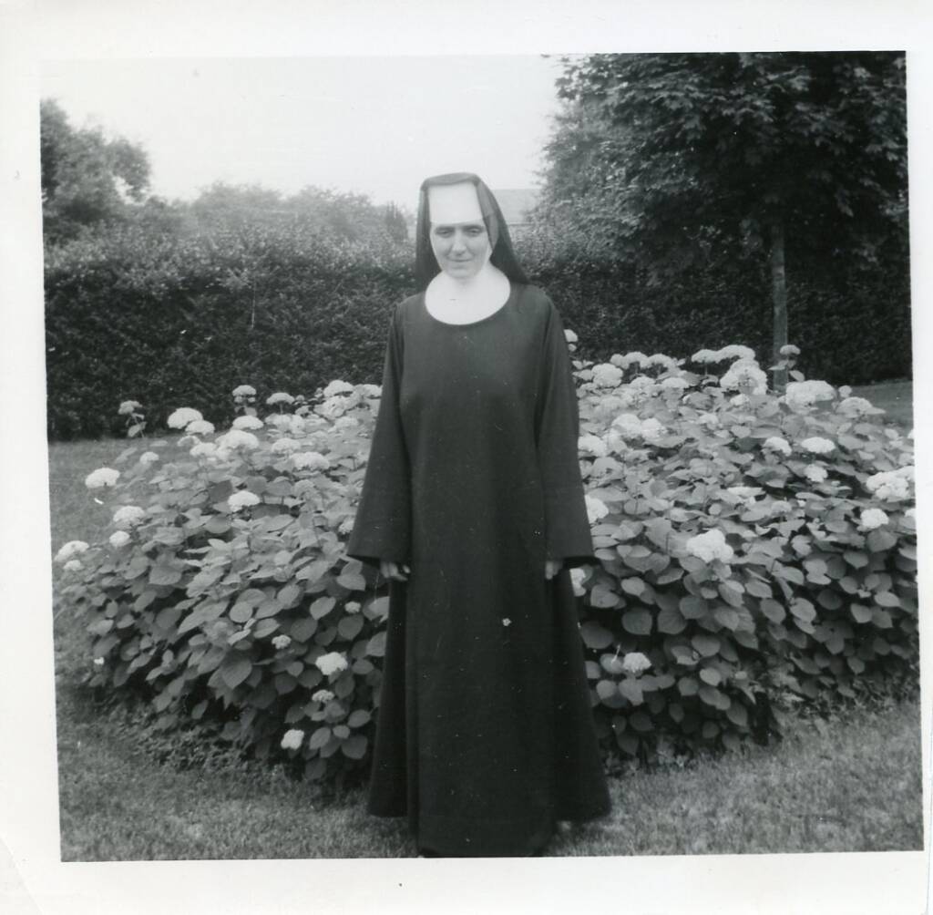 A nun stands in front of a bush of blooming flowers. She is wearing a traditional habit, including a veil. The background shows a well-kept garden with trees and a hedge. The photo is in black and white.