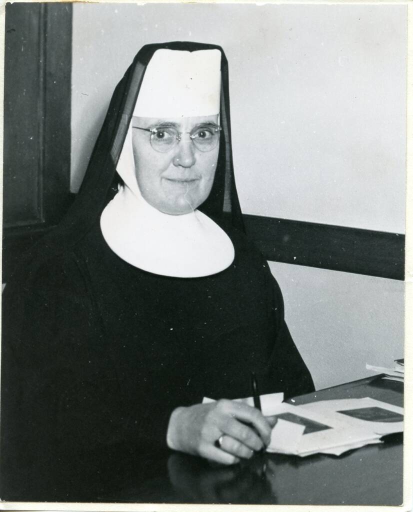 A nun wearing a traditional habit and glasses is seated at a desk, holding a pen and surrounded by papers. The background features a wooden panel wall.