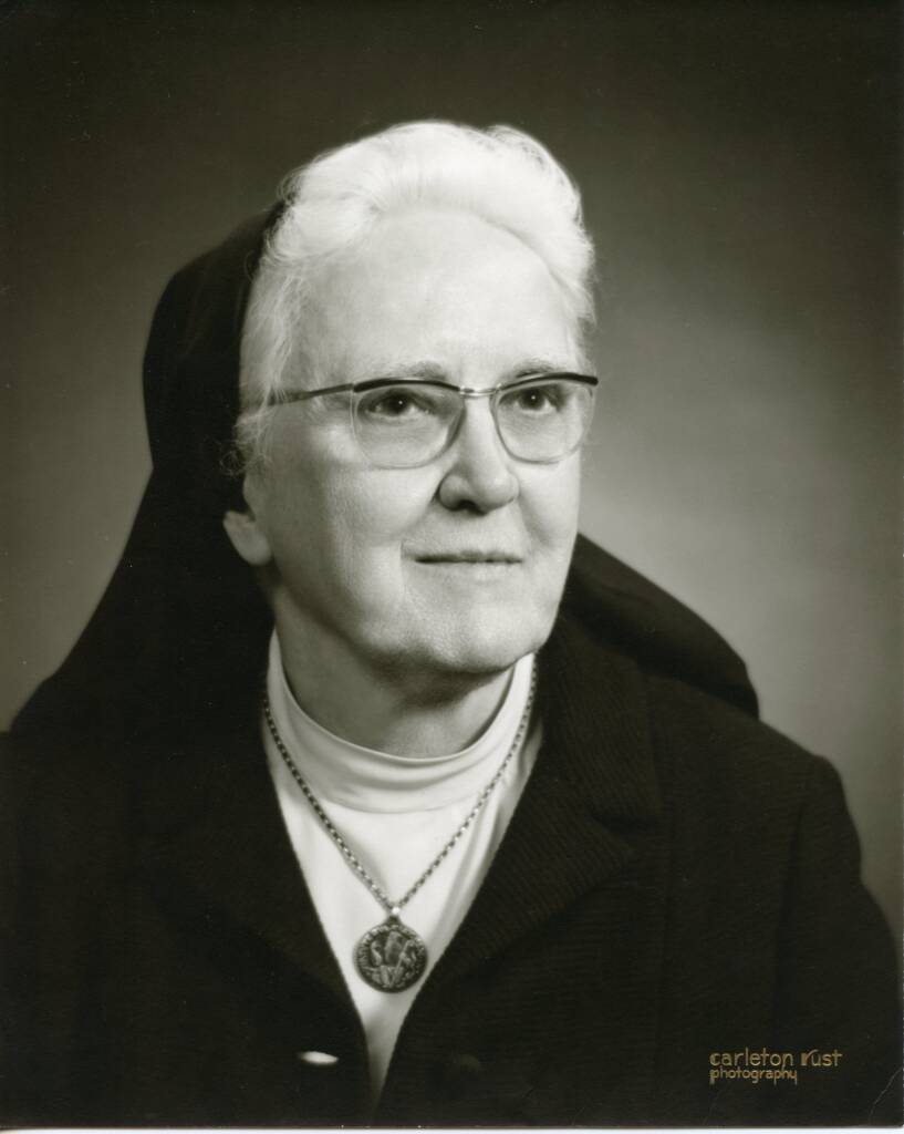 Black and white portrait of an elderly woman wearing glasses, a dark shawl over her head, and a necklace with a circular pendant. She gazes slightly to the side with a calm expression, against a plain background.