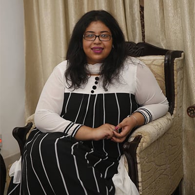 A person with long dark hair and glasses is sitting on a chair. They are wearing a black and white striped dress with buttons, and appear to be smiling. The background features beige curtains.