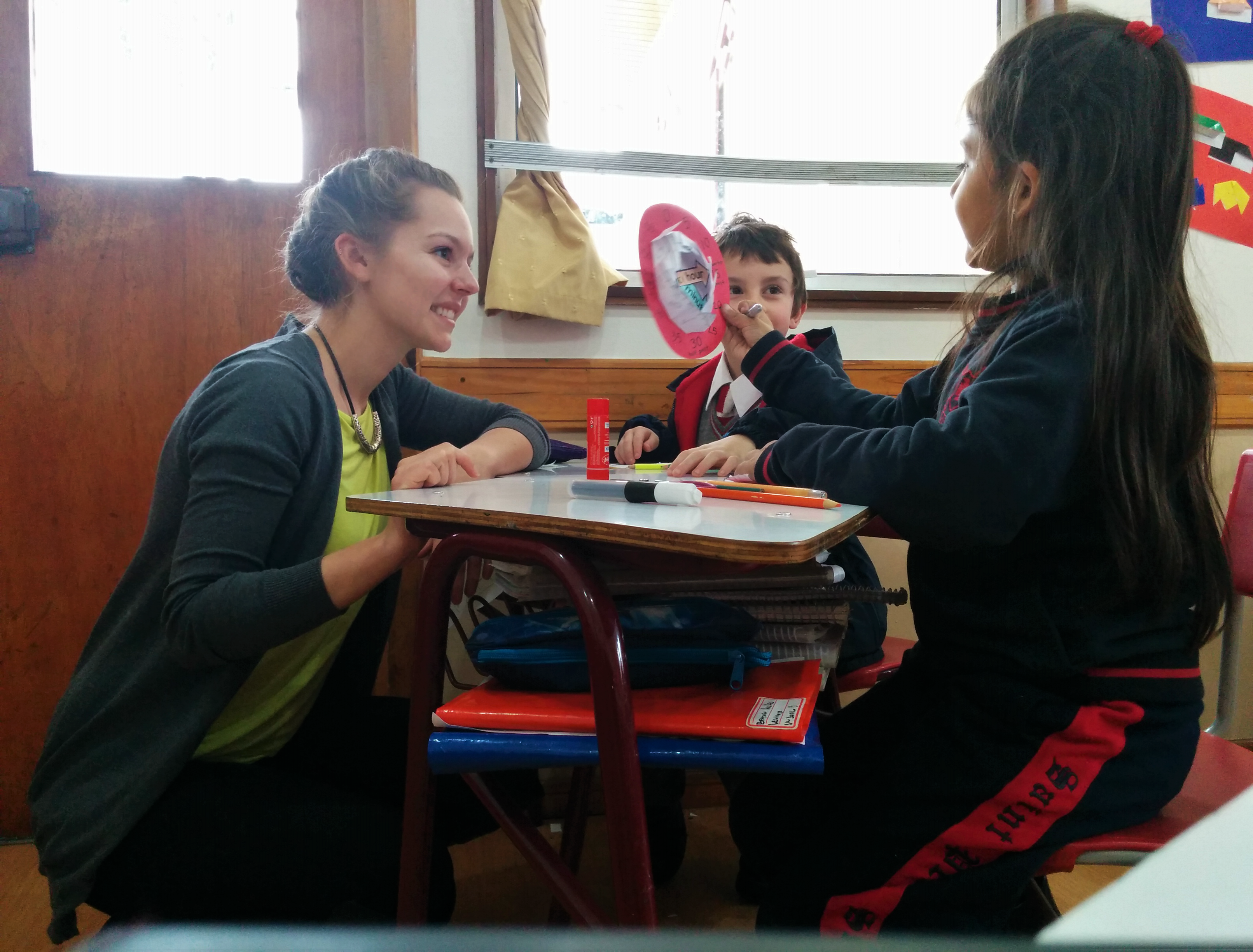A teacher kneels by a desk, smiling at two young children. One child holds a paper cutout and talks to the teacher, while both sit near a desk with school supplies. Classroom art decorates the walls in the background.