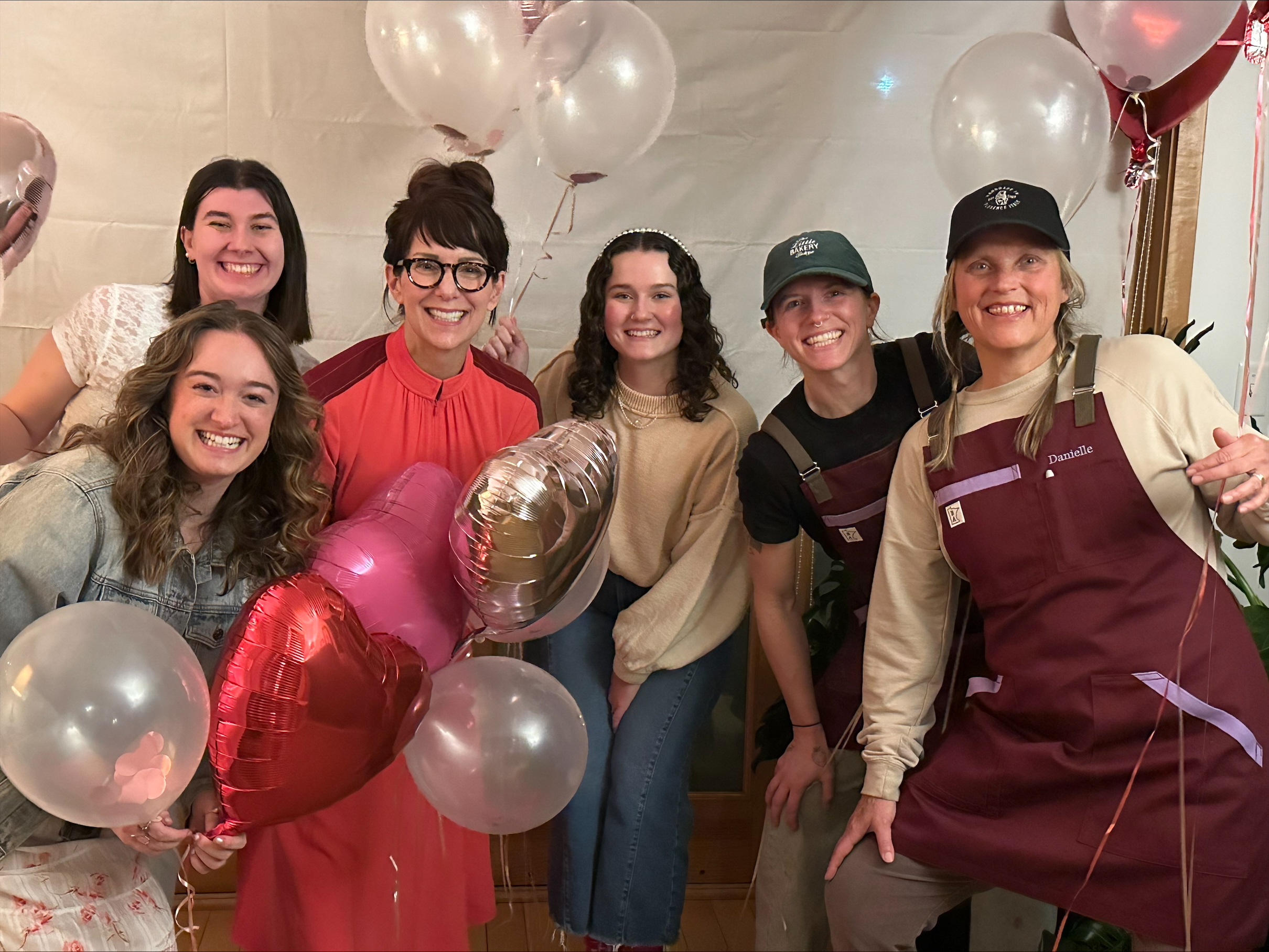 A group of six smiling people pose together, holding balloons in heart and round shapes. They are in casual and work attire, standing against a plain backdrop with potted plants visible. The atmosphere is festive and joyful.