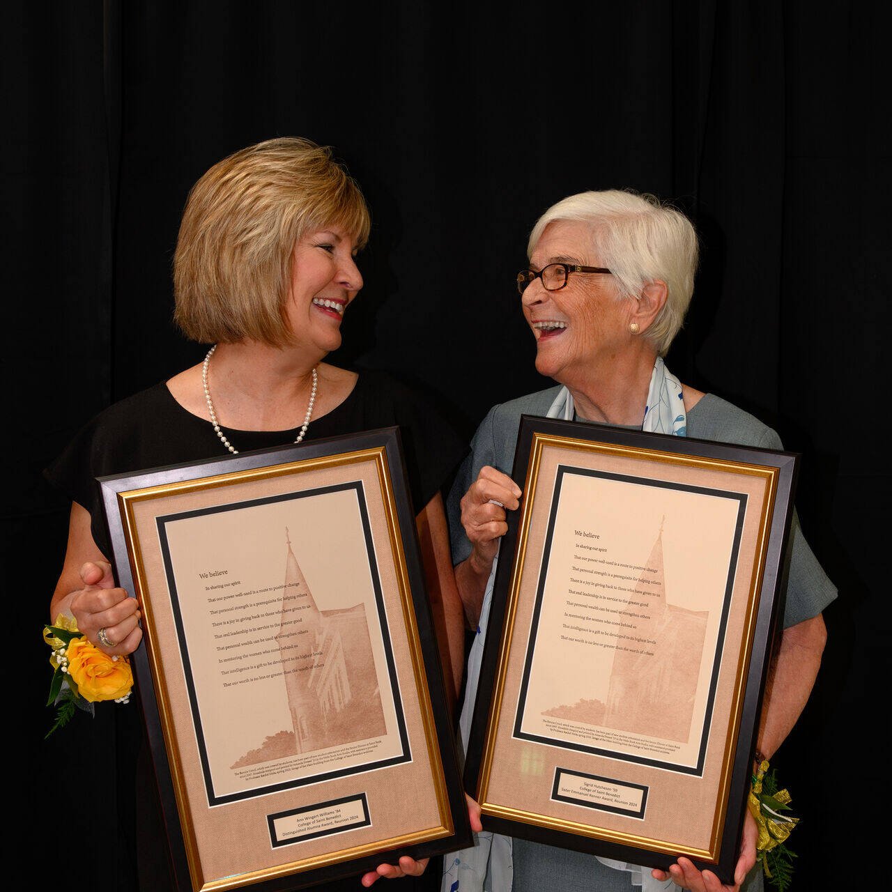 Two smiling women stand side by side, each holding a framed award and wearing yellow corsages on their wrists, with a black curtain as the background.