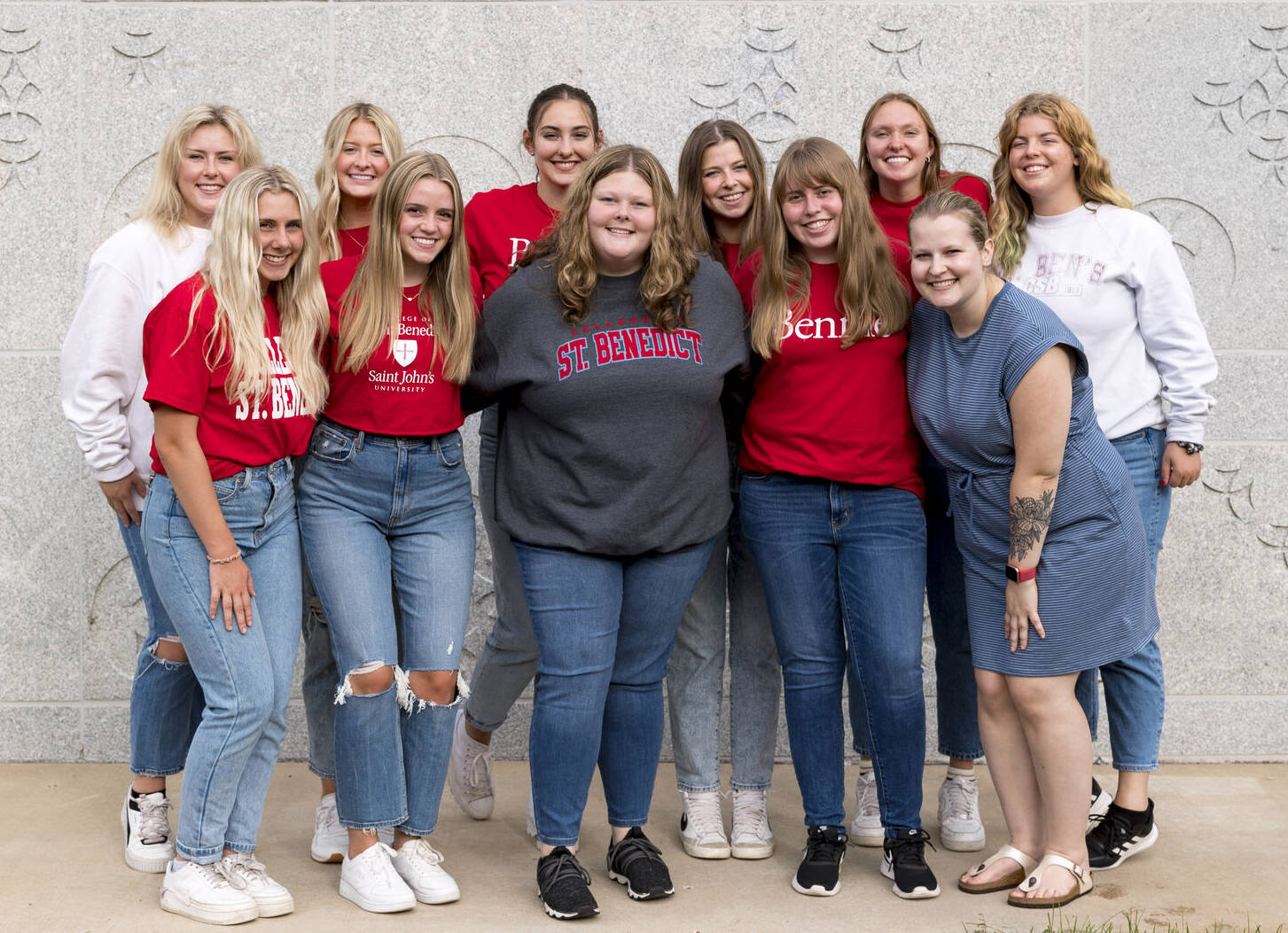 A group of twelve young women stand smiling in front of a gray stone wall. Most are wearing red, white, or gray St. Benedict shirts, and casual jeans. They appear happy and relaxed, posing together outdoors.