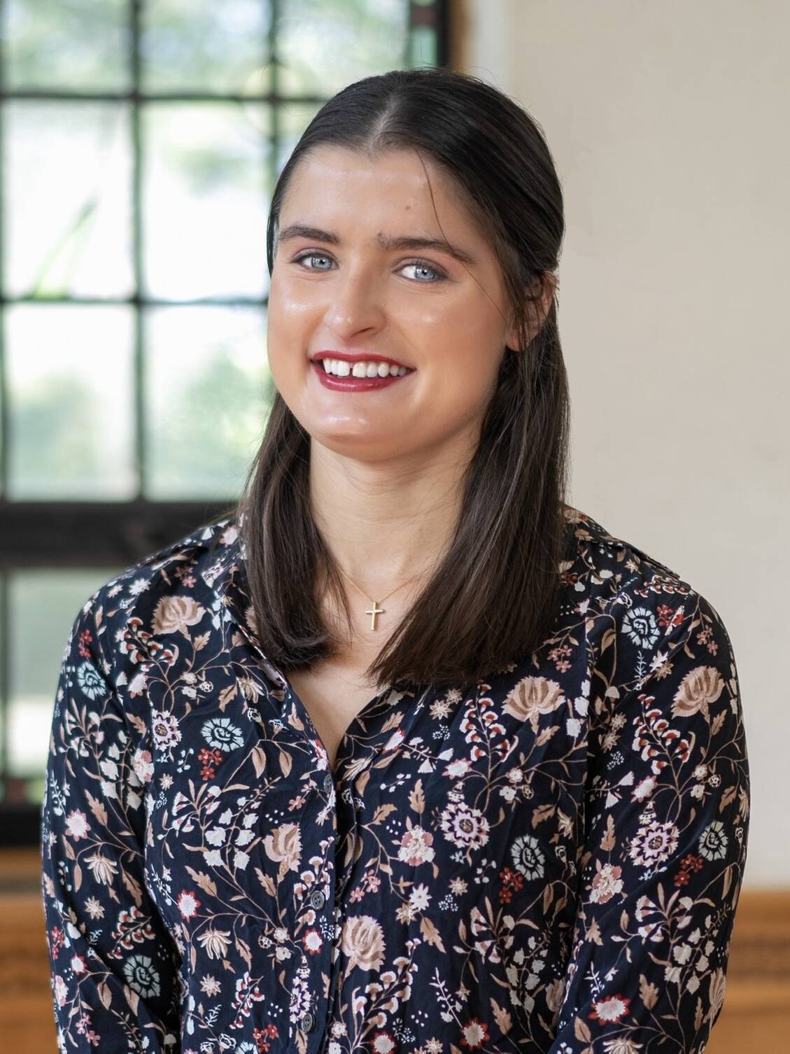 A woman with straight brown hair, wearing a dark floral blouse and a gold cross necklace, stands indoors in front of a window, smiling at the camera.