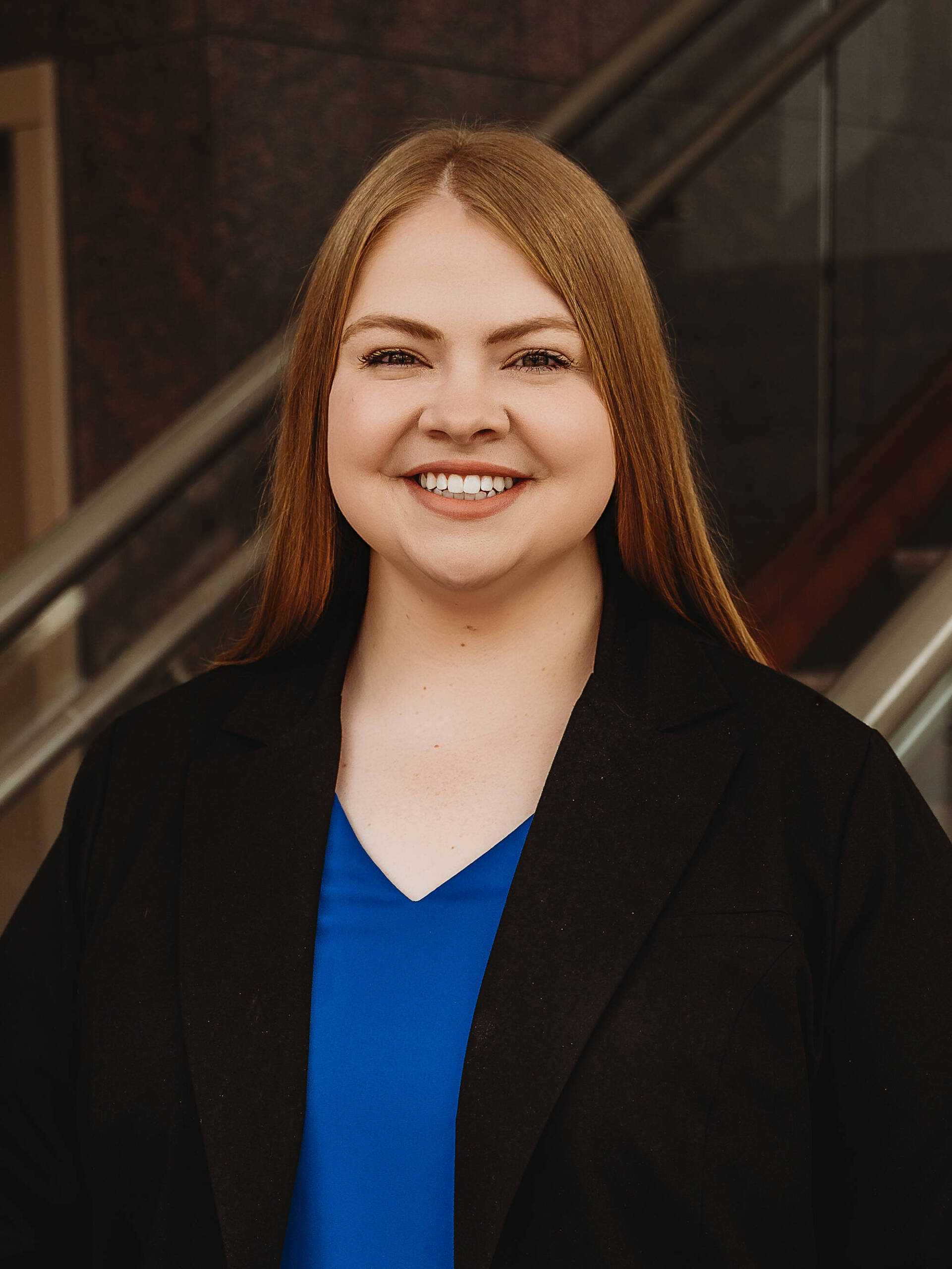 A woman with long straight hair, wearing a black blazer over a blue top, smiles while standing indoors in front of a staircase with glass and metal railings.
