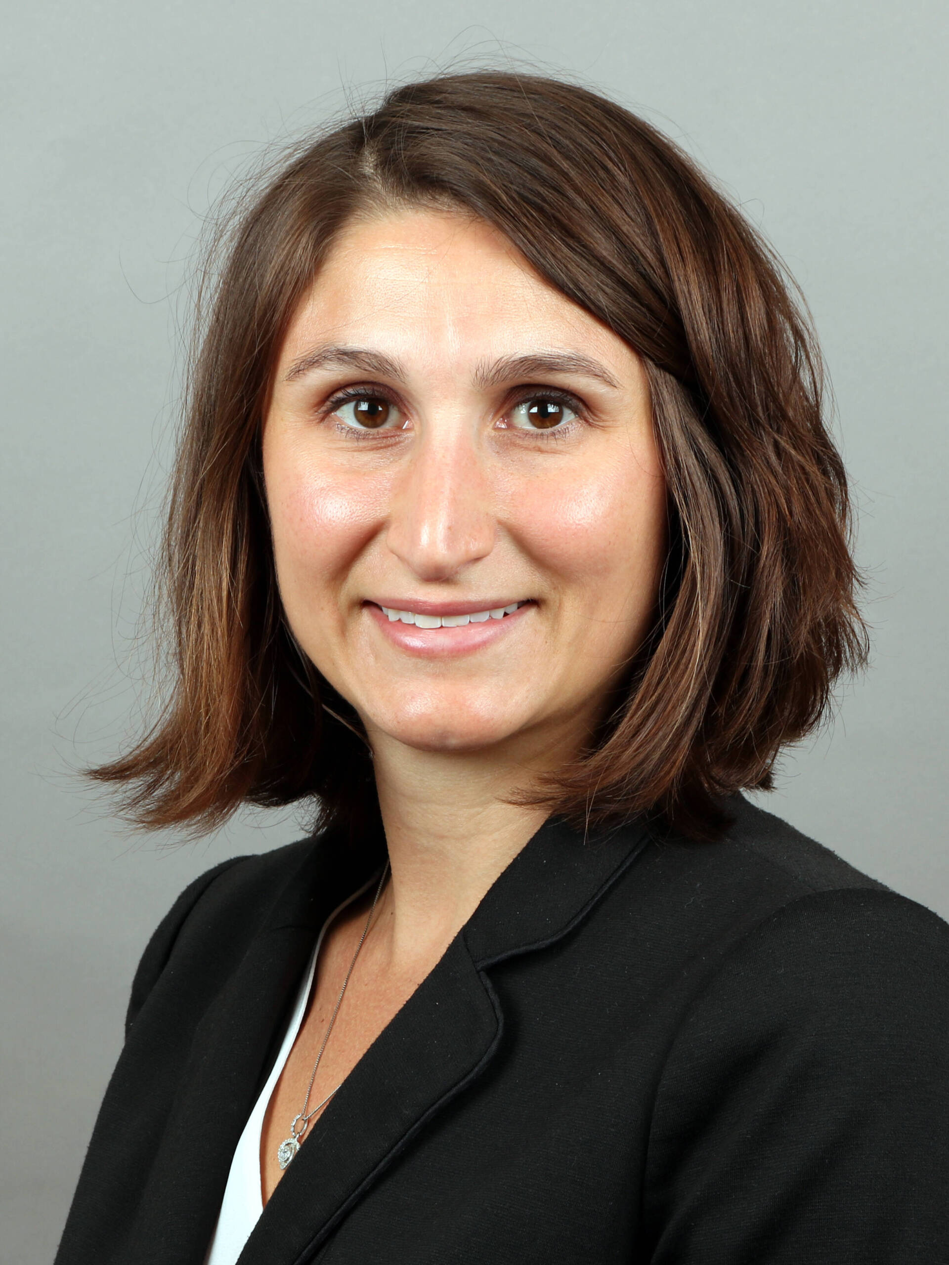 A woman with shoulder-length brown hair, wearing a black blazer and a white top, smiles at the camera against a gray background.