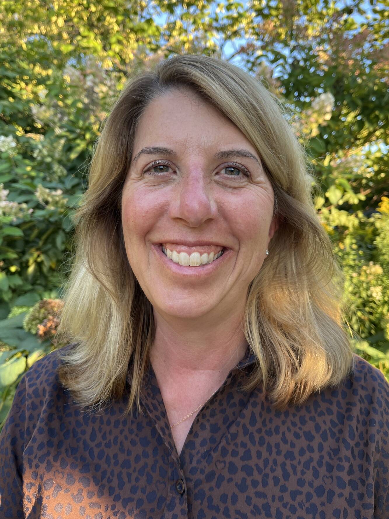 A woman with shoulder-length blonde hair smiles outdoors. She is wearing a dark, patterned blouse, and the background is filled with green foliage and sunlight.
