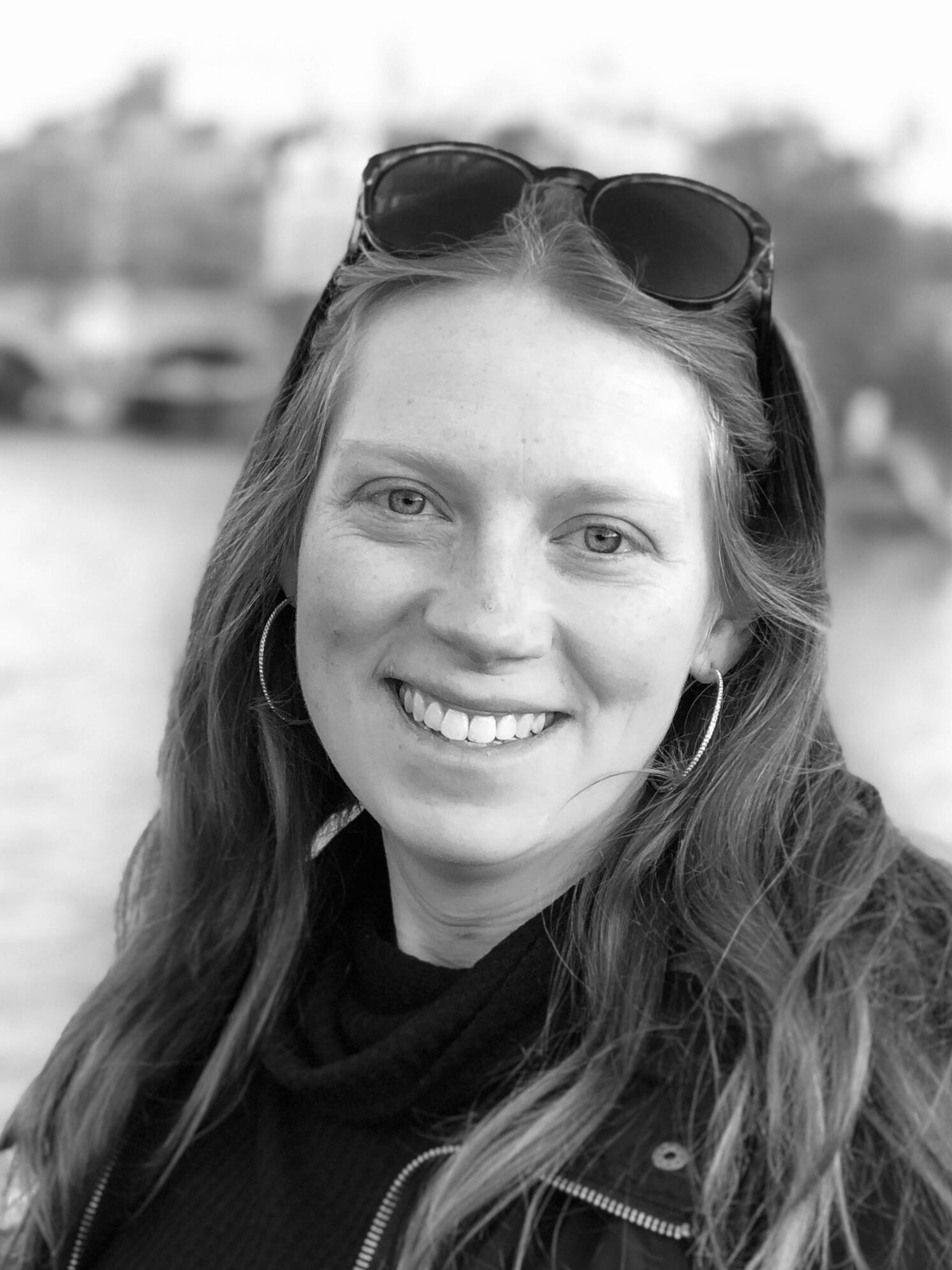 Black and white photo of a smiling woman with long hair, hoop earrings, and sunglasses resting on her head. She is wearing a black top with a high collar, standing outdoors with a blurred background showing a river and bridge.