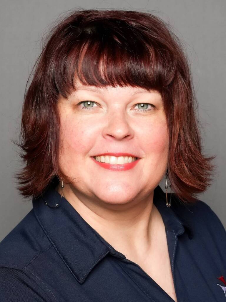 A person with shoulder-length brown hair and bangs, smiling at the camera. They are wearing a dark blue collared shirt and dangle earrings with a gray background.