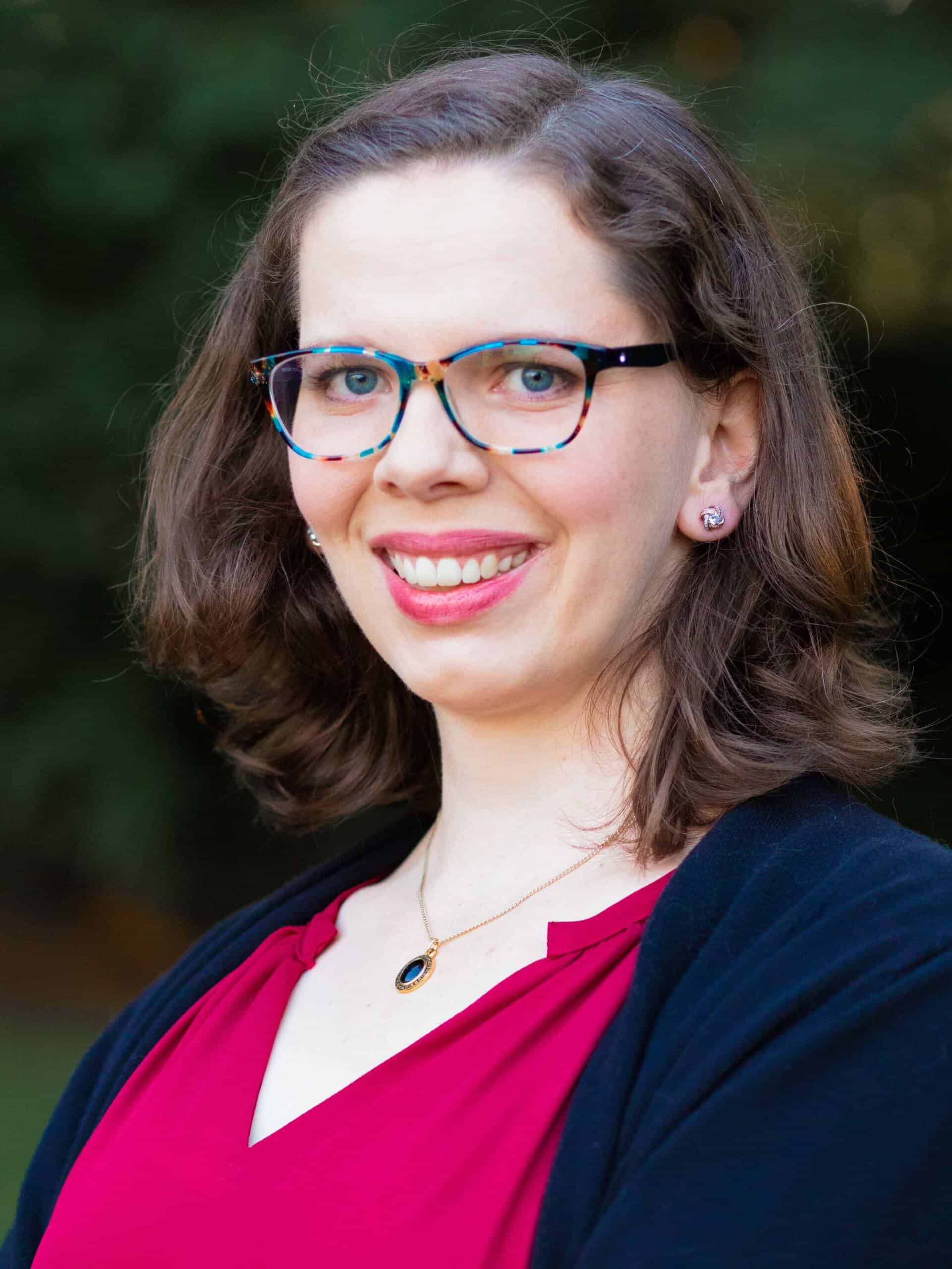 A woman with shoulder-length brown hair is smiling at the camera. She is wearing blue-rimmed glasses, a red top, a black cardigan, and a necklace with a dark pendant. The background is blurred, showing a natural outdoor setting.