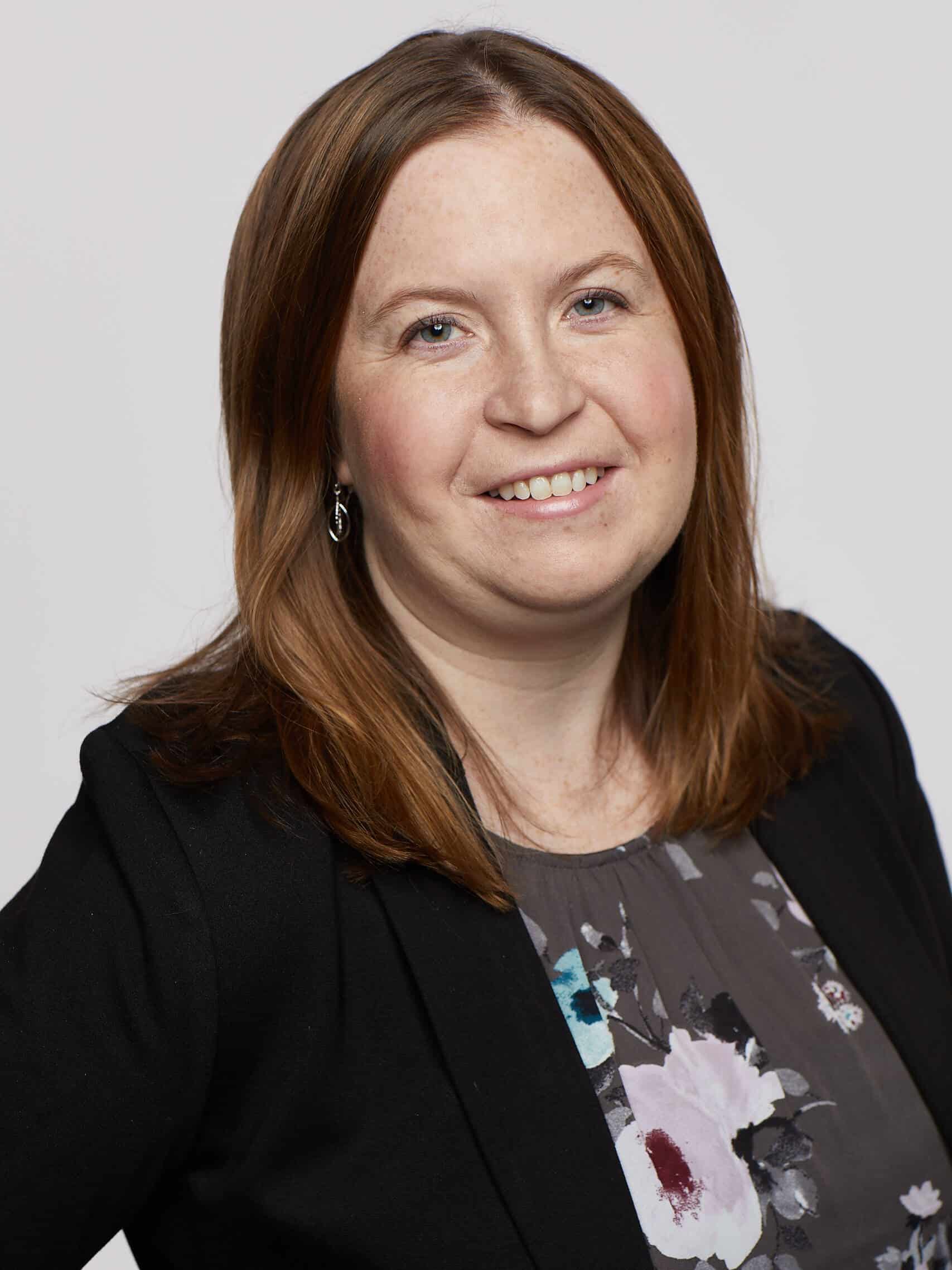 A woman with shoulder-length brown hair is smiling at the camera. She is wearing a black blazer over a floral blouse with shades of blue and purple flowers. The background is plain and light-colored.