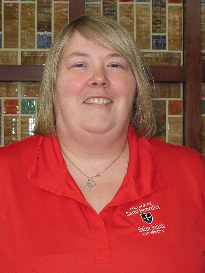 A person with short blonde hair is smiling. They are wearing a red polo shirt with the logos of the College of Saint Benedict and Saint John's University. The background features a colorful mosaic tile wall.