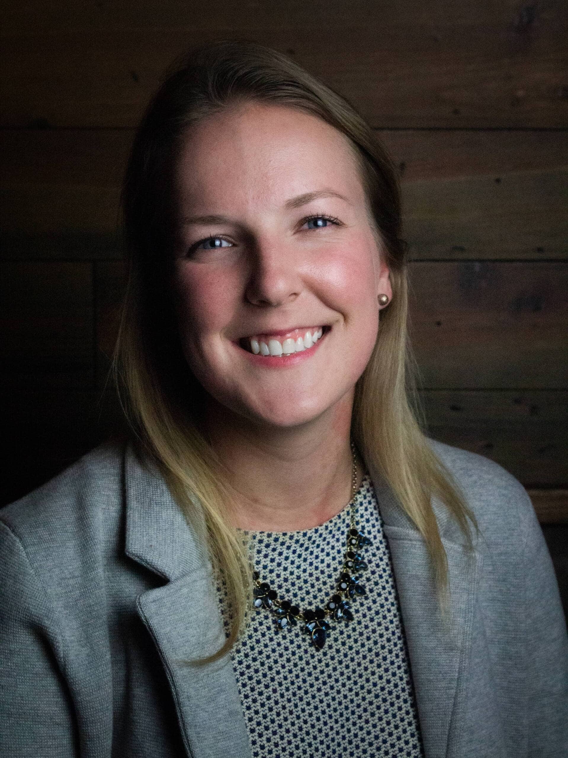 A person with long blonde hair, smiling, wearing a light gray blazer over a patterned blouse, and a dark beaded necklace. The background consists of wooden panels in various shades of brown.