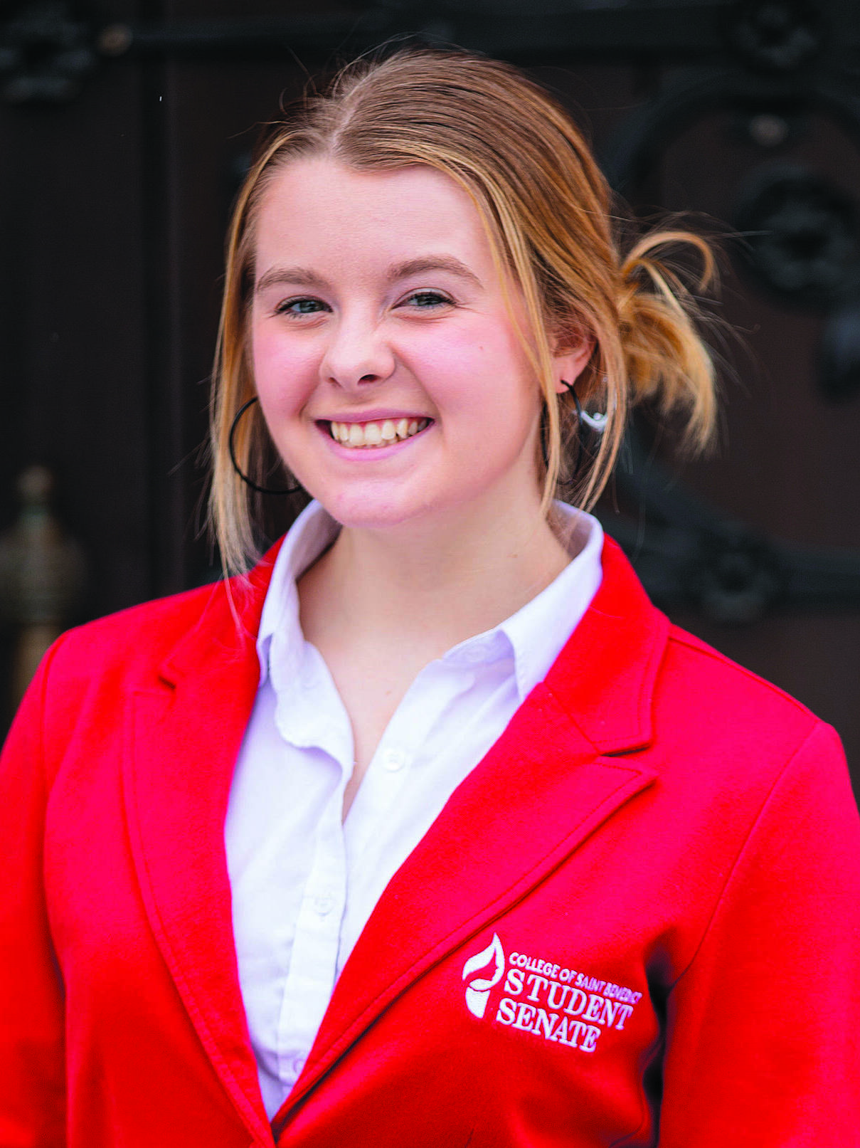 A young woman with blonde hair tied back is smiling at the camera. She is wearing a white blouse and a red blazer with an emblem on the left side that reads "Student Senate." The background is slightly blurred with a dark, intricate design.