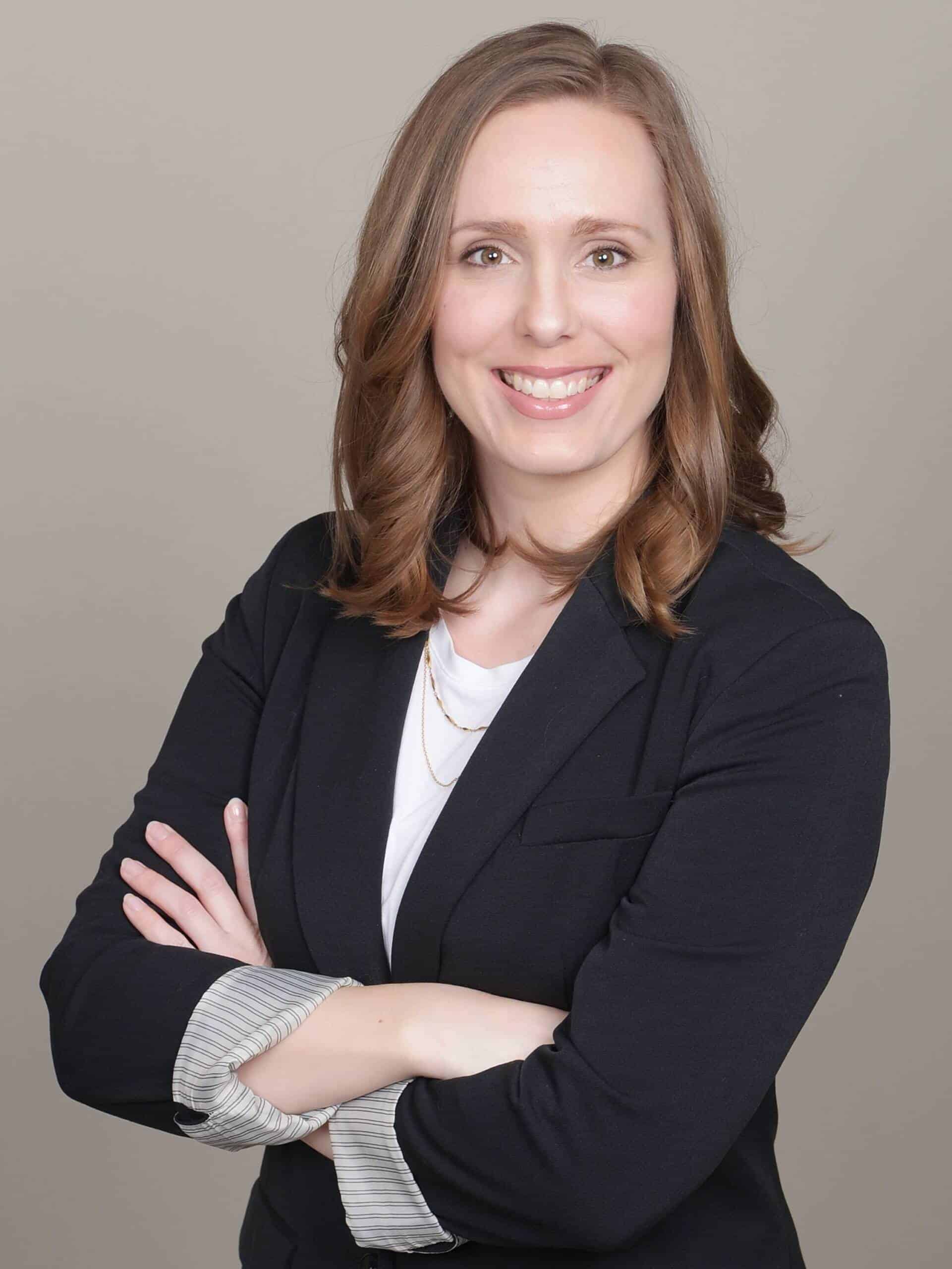 A woman with shoulder-length brown hair is smiling and posing with her arms crossed. She is wearing a black blazer over a white top, and the background is a plain, light color.