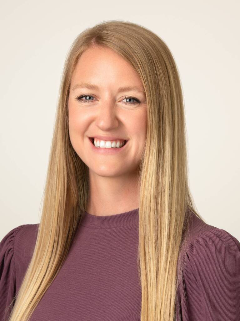 A smiling woman with long, straight blonde hair is shown in a portrait against a light background. She is wearing a mauve-colored top with puffed sleeves.