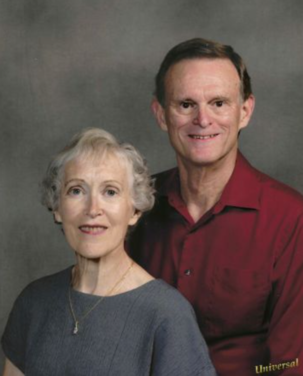 An older woman with short gray hair and a gray top sits in front of a smiling older man with short brown hair and a maroon shirt, posing together against a neutral gray background.