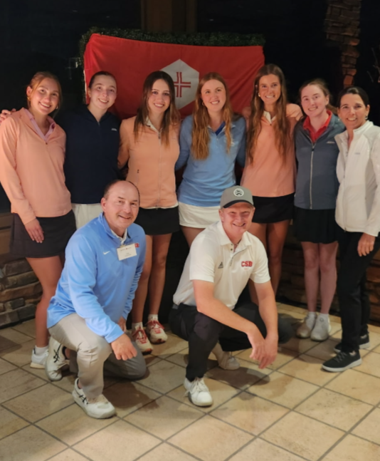 A group of eight people, six women standing and two men kneeling in front, pose indoors on a tiled floor with a red banner featuring a white logo in the background. Everyone is smiling and dressed in sporty attire.