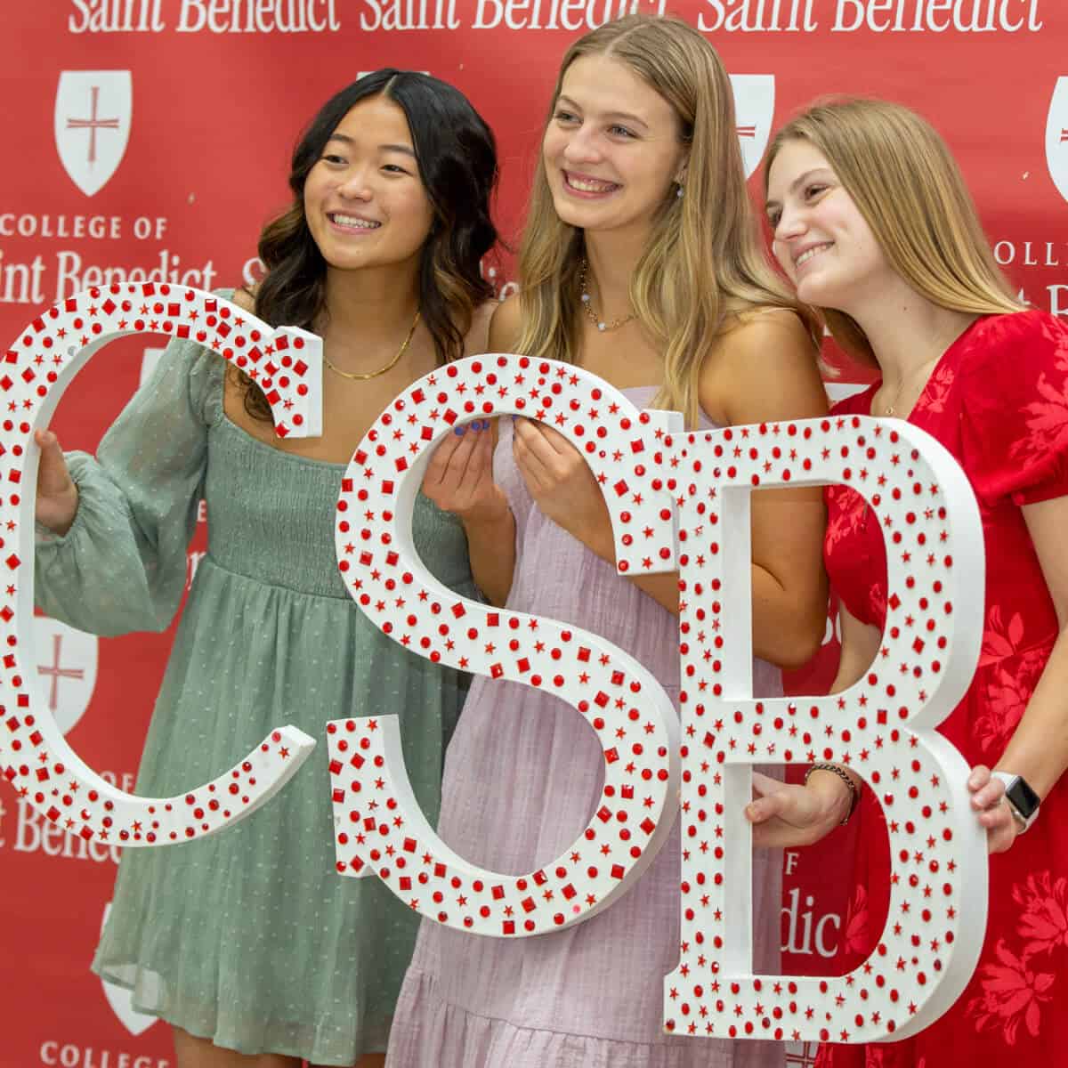 Three young women smile and pose together at a photo booth, holding a large "CSB" sign. A red backdrop behind them features the logo and name of the College of Saint Benedict.