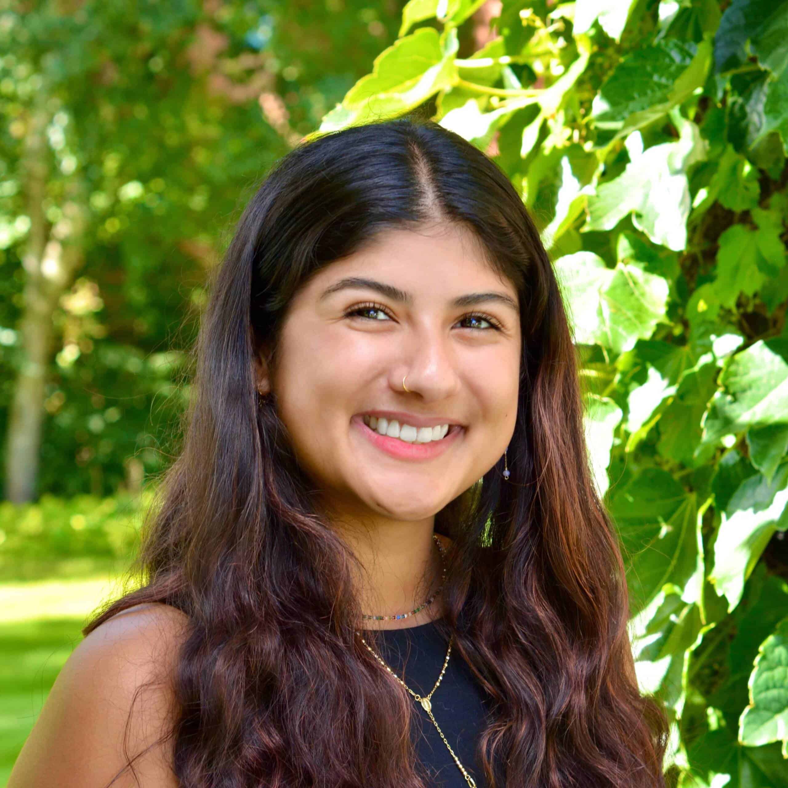 A young woman with long, wavy brown hair smiles while standing outdoors in front of lush green ivy leaves and sunlight filtering through trees. She is wearing a sleeveless black top and layered necklaces.