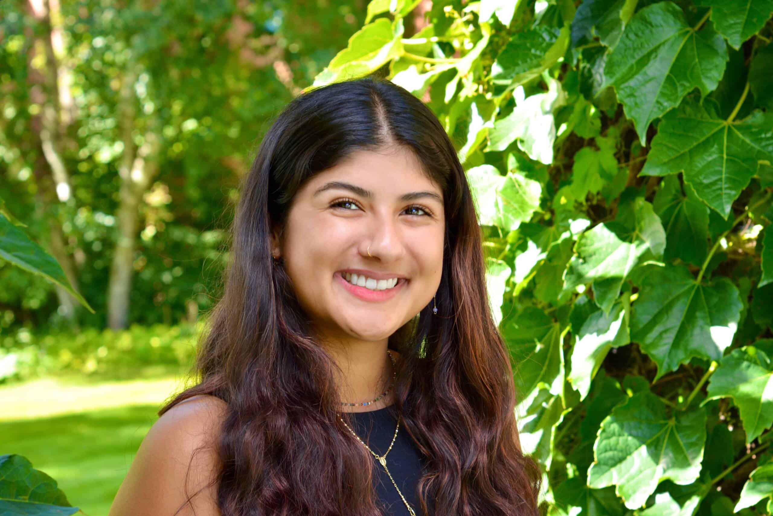 A young woman with long, wavy brown hair smiles while standing outdoors in front of lush green ivy leaves and sunlight filtering through trees. She is wearing a sleeveless black top and layered necklaces.