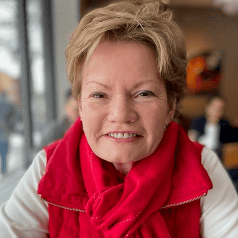 A smiling older woman with short light brown hair wearing a bright red scarf and vest sits indoors by a window, with a blurred background of people and soft lighting.