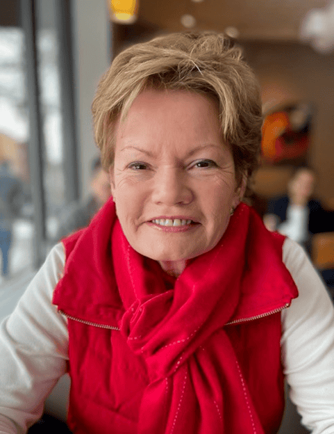 A smiling older woman with short light brown hair wearing a bright red scarf and vest sits indoors by a window, with a blurred background of people and soft lighting.