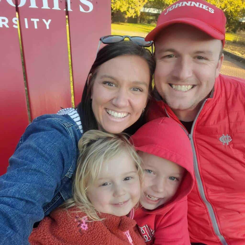 A smiling family of four poses closely together outdoors, with two children in red jackets in front of a woman and a man wearing a red "Johnnies" cap. A red sign reading "Benedict St. John's University" is partially visible behind them.
