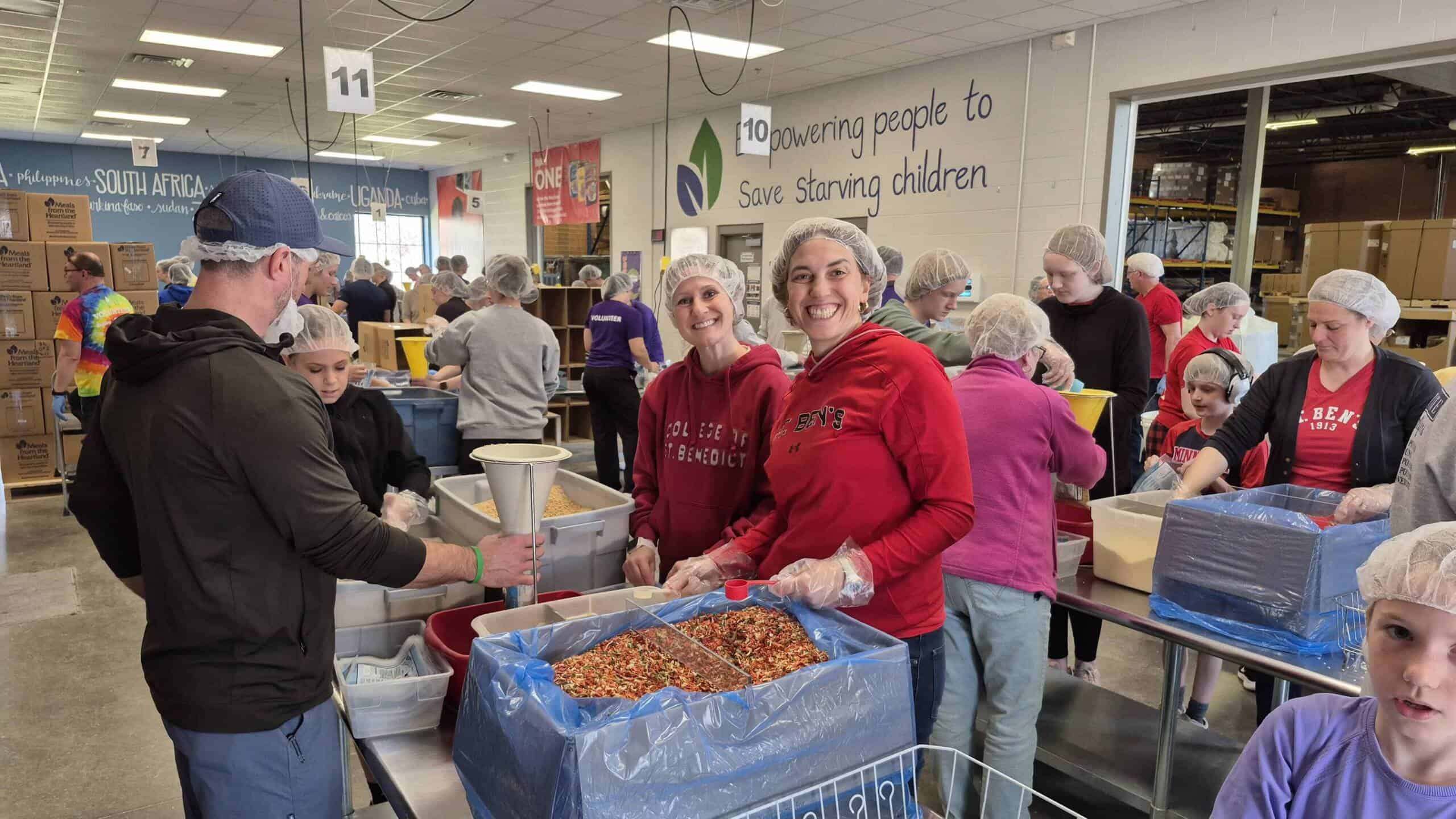 Volunteers wearing hairnets and gloves pack food into bags at an indoor facility. Large bins of ingredients are on tables, and people of various ages work together. A sign on the wall reads, “Empowering people to save starving children.”.