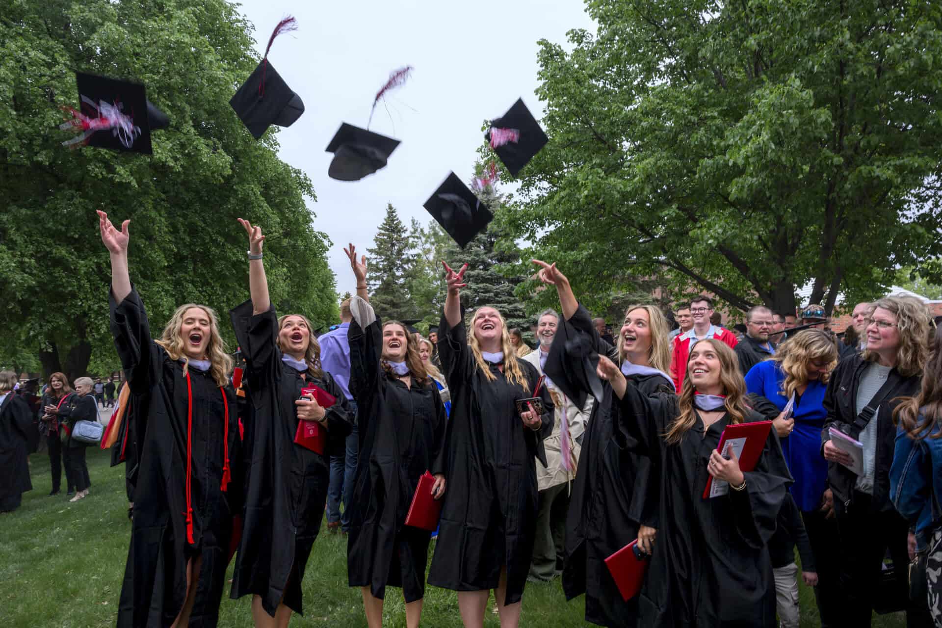 A group of graduates in black gowns joyfully throw their caps into the air outdoors, surrounded by friends and family who watch and celebrate amid green trees.