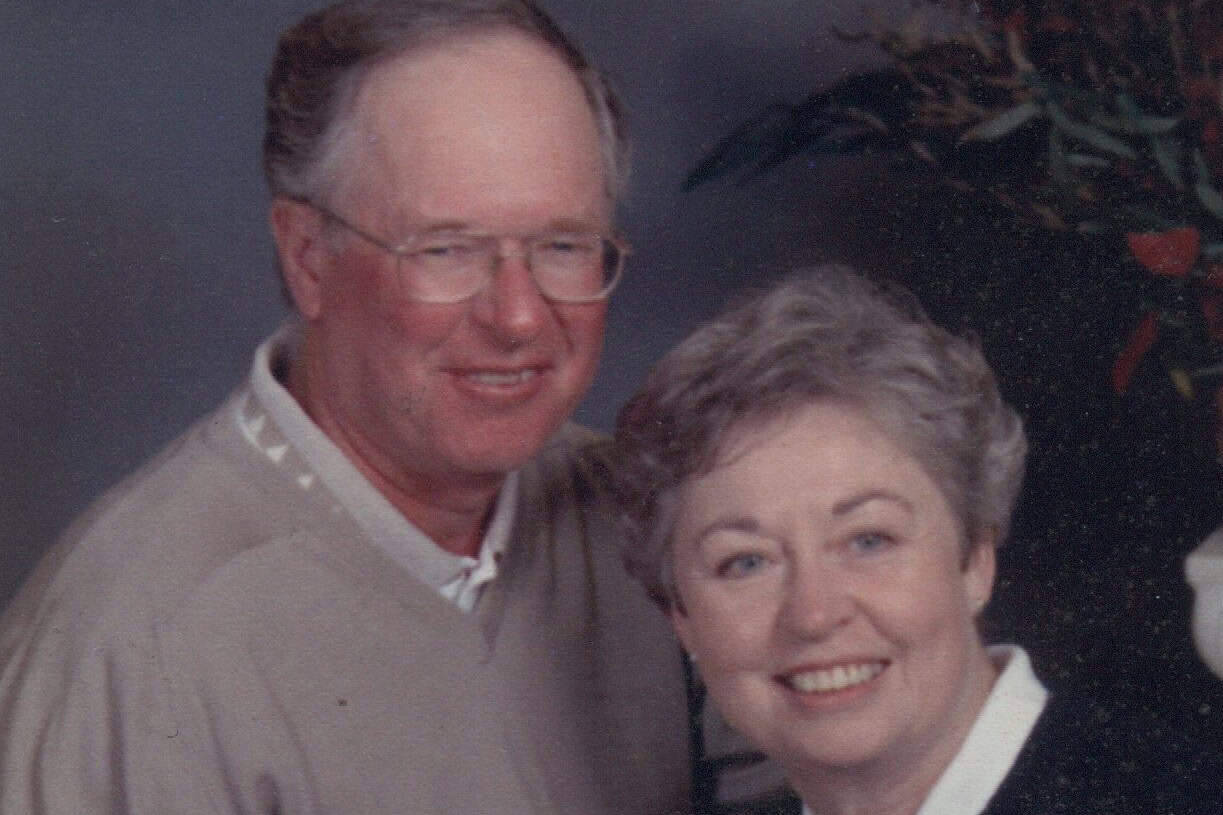 An older man and woman are sitting close together, smiling at the camera. The man wears glasses and a beige sweater, while the woman has short gray hair and wears a black sweater over a white shirt. A floral arrangement is in the background.